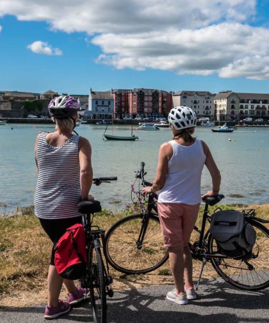 Two cyclists on a Cycling Tour taking a break to enjoy the view of a picturesque harbor dotted with boats, under a blue sky with fluffy clouds.