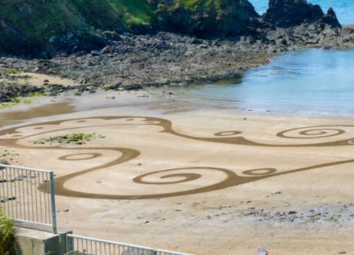 A tranquil Copper Coast beach scene with artistic sand patterns, a single sunbather at the corner, and serene waters bordered by rocky shores.