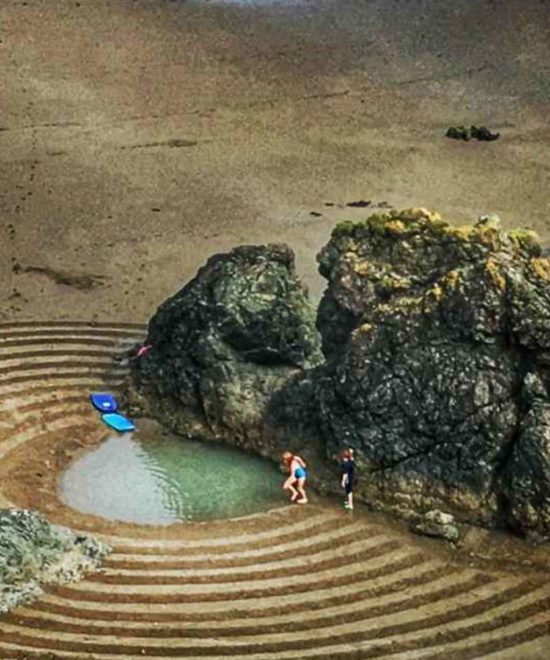 Aerial view of two people standing near a natural tidal pool on the Copper Coast, surrounded by dark rocks and patterned sand art on a beach, with a blue surfboard resting on the sand.