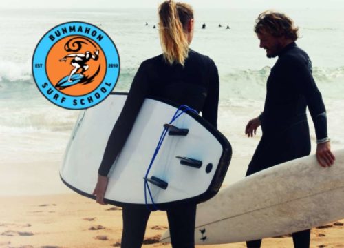 Two surfers, one holding a surfboard, standing on the beach with gentle waves in the background, possibly discussing surfing techniques with a logo for Bunmahon Surf School visible in the upper left corner