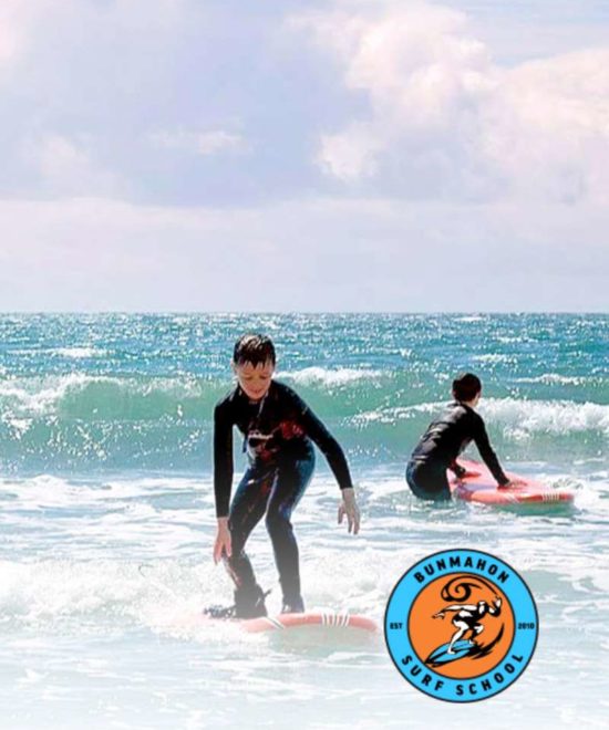 Two individuals in wetsuits surfing on surfboards along the Copper Coast, under a clear blue sky and ocean in the background, promoting the experience at a surf school.