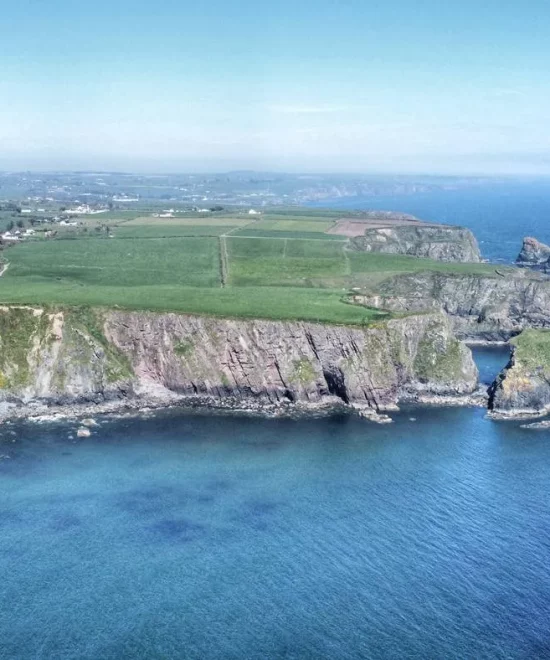 A scenic aerial view of Ballydwan Bay features steep cliffs, rocky formations, and clear blue waters. The coastline is bordered by green fields and farmland, extending into the distance. The sky is clear with a few clouds, adding to the tranquil beauty of this picturesque coastal landscape.