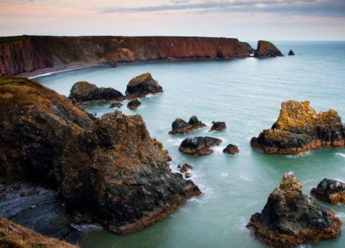 Rugged cliffs and scattered sea stacks overlook a tranquil cove at twilight, displaying the serene yet dramatic landscape of Ballydwan Bay, Ireland.