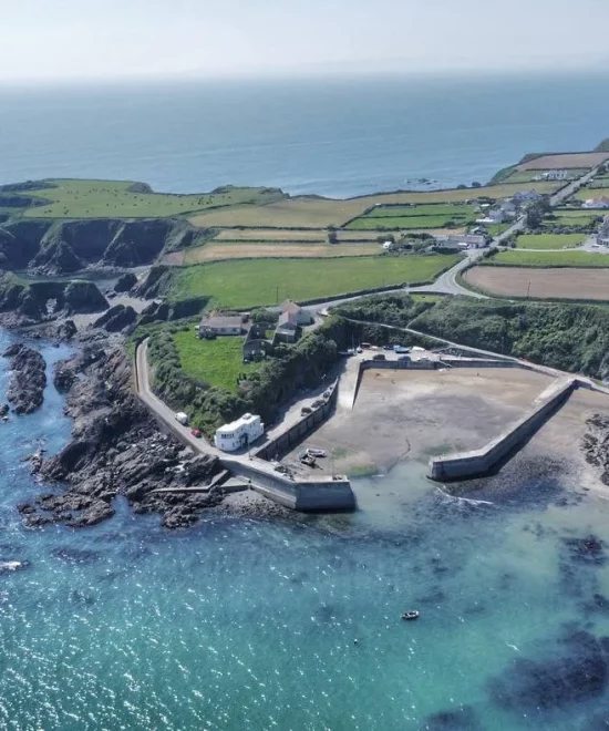 Aerial view of the coastal landscape near Dunabrattin Head, featuring a small port at Boatstrand Harbour with breakwater structures, surrounded by blue waters and rocky shores. Green fields and rural buildings are visible on cliffs above the coast, extending into the horizon where the blue sky meets the sea.