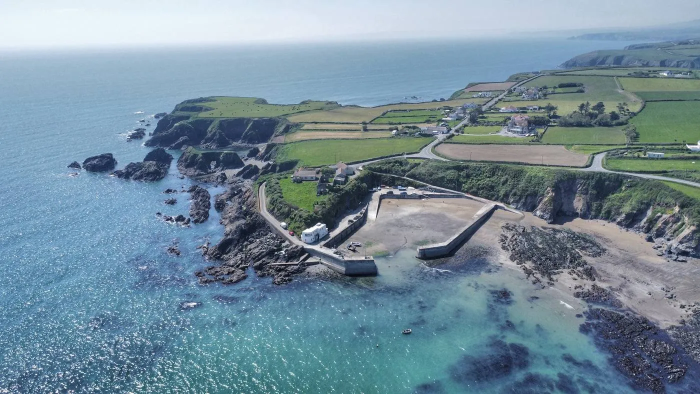 Aerial view of the coastal landscape near Dunabrattin Head, featuring a small port at Boatstrand Harbour with breakwater structures, surrounded by blue waters and rocky shores. Green fields and rural buildings are visible on cliffs above the coast, extending into the horizon where the blue sky meets the sea.