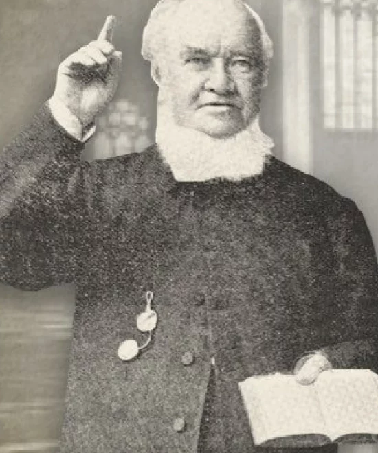 A black-and-white photograph of David Alfred Doudney, an Evangelical clergyman dressed in formal 19th-century attire, holding a small open book in one hand and pointing upwards with the other. He has a stern expression and appears to be giving a speech or lecture, with a church interior in the background.