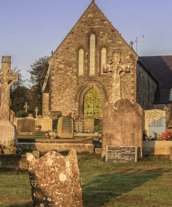 A historic stone church with arched windows, known as Dunhill Old Church, stands surrounded by a graveyard filled with various tombstones and crosses. The sunlight casts a warm glow, highlighting the weathered gravestones and the church's intricate stonework. The grass surrounding the graves is lush and green.