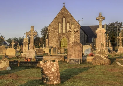 A historic stone church, known as Dunhill Old Church, stands among weathered gravestones and monuments in a serene graveyard. The early morning light casts a warm glow on the scene, highlighting the ancient architecture and lush green grass.