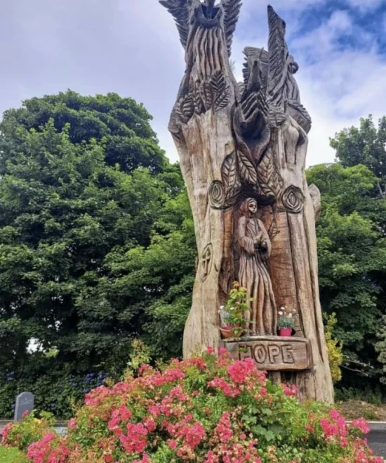 A large, intricately carved wooden statue stands among vibrant pink flowers and lush green trees. The carving depicts various figures and symbols, including an eagle at the top and a figure of a saint or religious person in the center. The word "HOPE" is carved at the base, echoing Fenor's rich history.