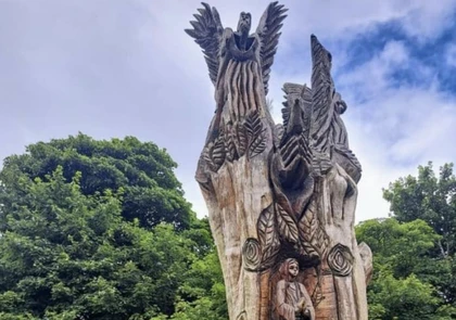 A large wooden sculpture stands in front of a lush green background in Fenor. The piece, rich with history, features a variety of carved figures, including wings at the top, cascading details, and a central human-like figure at the base. The sky is partly cloudy with patches of blue.