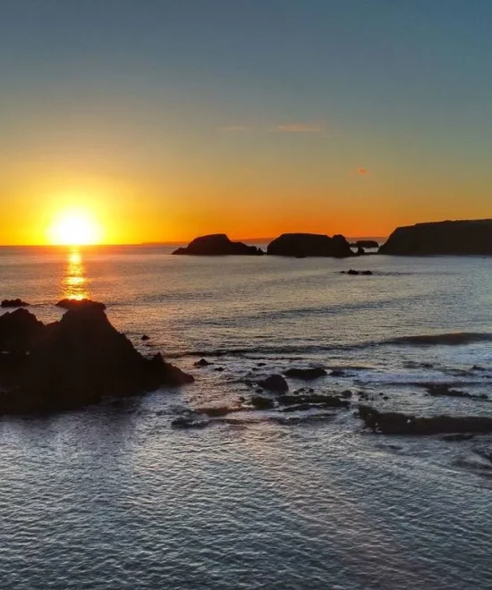 A breathtaking sunset over Garrarus Strand in Ireland with the sun partially hidden behind a distant line of jagged coastal cliffs. The water shimmers with orange and gold hues, and several large rocks and small waves are visible in the foreground. The sky transitions from blue to orange.