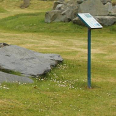 An information sign next to a rock in a grassy area of the Geological Garden, possibly providing details about the geology or history of the location.