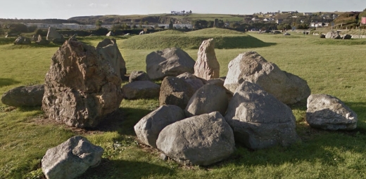 A collection of large, irregularly shaped rocks arranged in a geological formation on a grassy field with distant hills and scattered buildings in the background under a clear sky.