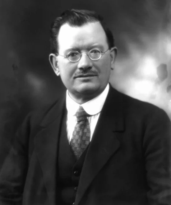 Black-and-white portrait of John Wheatley, a Labour MP, wearing round glasses, a suit, and a tie. He has neatly combed hair and a slight smile. The background is blurred, directing focus on Wheatley's face and attire.