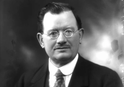 Black and white portrait of Labour MP John Wheatley, a middle-aged man with short, neatly combed hair, wearing round glasses, a dark suit, white shirt, and patterned tie. He has a distinct mustache and is posing against a blurred, neutral background.