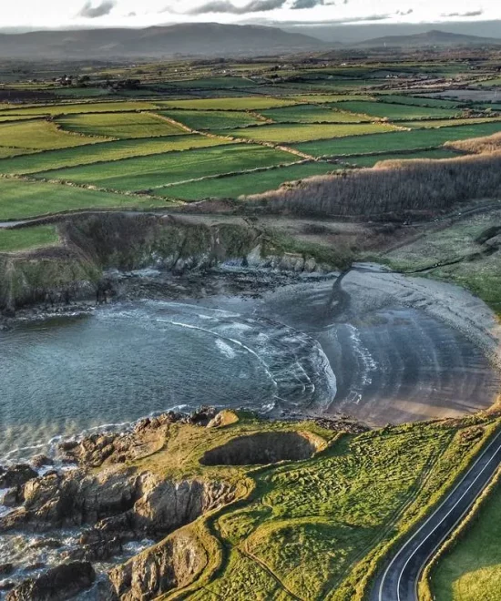 Aerial view of Kilmurrin Cove, a coastal landscape featuring a serene cove with sandy and rocky shores, surrounded by green fields and farmlands. A winding road runs parallel to the coastline, and rolling hills are visible in the distance under a partly cloudy sky.