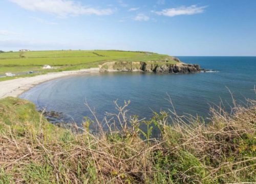 A serene coastal landscape featuring Kilmurrin Cove with a curving sandy beach, surrounded by grassy headlands under a clear blue sky.