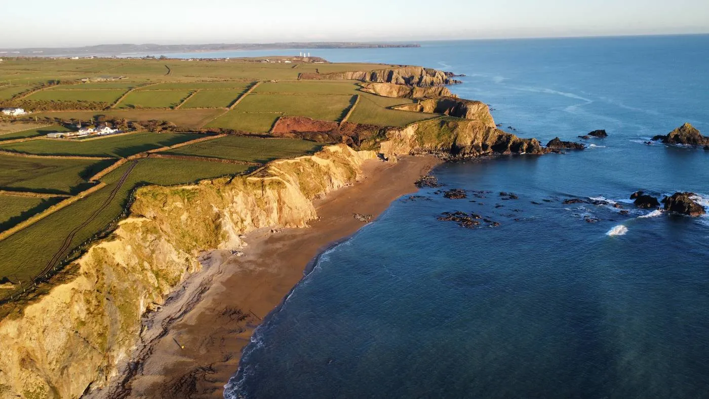 Aerial view of rugged coastal cliffs with green fields atop, leading to a sandy beach and rocky shoreline. The ocean waves gently meet the beach, and the coastline stretches into the distance under a clear sky at Garrarus Strand.