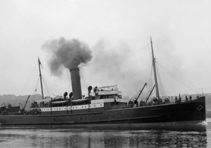 A black and white photograph showcases a vintage 19th-century steamship, emitting smoke from its central funnel. The vessel is on calm waters, with some crew members visible on deck. The shoreline is faintly visible in the background, under an overcast sky.