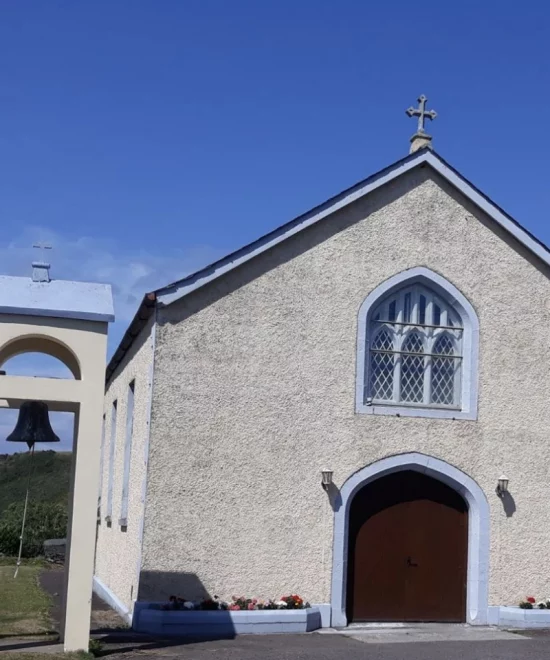 A small, beige chapel, reminiscent of St. Mary's Church in Saleen, with a wooden door and arched window stands under a clear blue sky. To the left, a bell hangs in a simple open frame. The chapel is adorned with potted flowers along its base. A green hill is visible in the background.