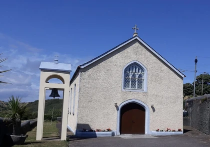 A small, cream-colored chapel, known as St. Mary's Church, with an arched doorway and a cross on top stands under a clear blue sky. To the left, there's a standalone bell tower with a single bell. Small plants and flowers line the base of the chapel's front wall in Bonmahon.