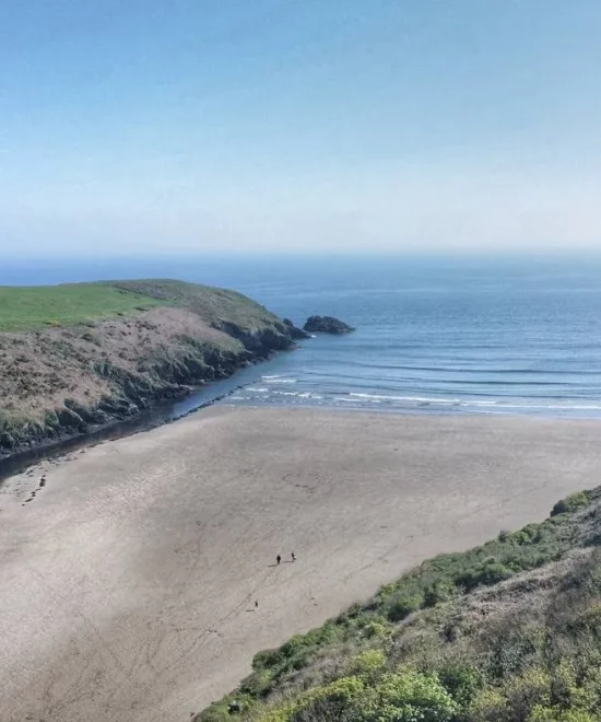 A serene coastal scene featuring Stradbally Cove with a sandy beach where gentle waves meet the shore. Green cliffs and grassy hills frame the beach. A small stream winds its way towards the sea, and two tiny figures can be seen walking on the sand under a clear, blue sky.