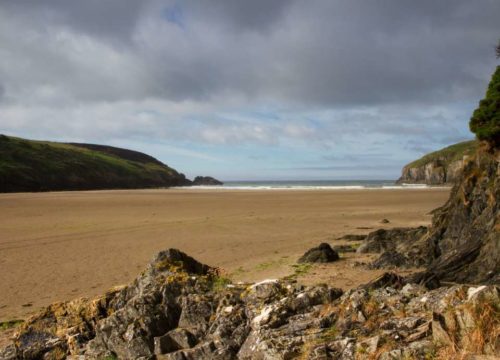 A tranquil sandy beach at Stradbally Cove, bordered by rugged cliffs under a partly cloudy sky, with the calm ocean in the distance.