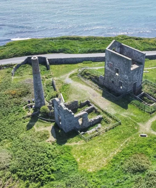 Aerial view of Tankardstown's coastal landscape featuring historic ruins, including a large, roofless stone building and a tall, narrow chimney. The ruins are surrounded by green grass and situated near a winding road. The sea is visible in the background.