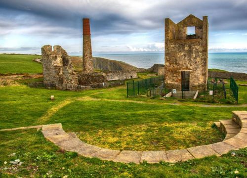 The ruins of an industrial past: a dilapidated structure with a tall chimney stands against a backdrop of lush coastal scenery at Tankardstown House, remnants of a bygone era where nature slowly re