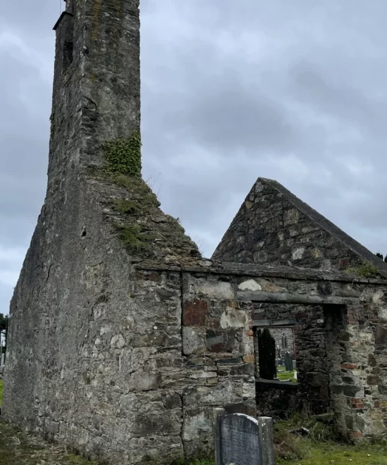 An old, weathered stone church stands partially in ruins under a cloudy sky. Ivy climbs part of the tall, narrow steeple. In the foreground, there is a gravestone with some flowers placed at its base. The scene appears somber and historical, perfect for travel enthusiasts exploring quaint villages.