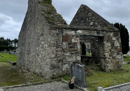 A weathered, stone church ruin stands amid a graveyard in the Village of Kill. The structure has partially collapsed walls with some portions still standing. The surrounding area has headstones and greenery, with a cloudy sky overhead.