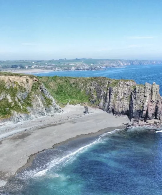 A scenic coastal landscape featuring tall rocky cliffs at Bunmahon Head, a stretch of sandy and pebbly beach known as Tra na mBo, and clear blue ocean water. Green vegetation covers parts of the cliffs. In the distance, the coastline extends out with more cliffs and land visible under a clear blue sky.