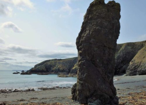 A solitary rock formation stands on Tra na mBo, with a calm sea and Bunmahon Head in the background, under a partly cloudy sky.