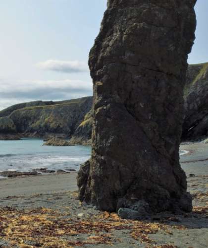 A towering rock formation stands guard on Tra na mBo, with gentle waves lapping at the shore and a backdrop of serene coastal cliffs at Bunmahon Head.