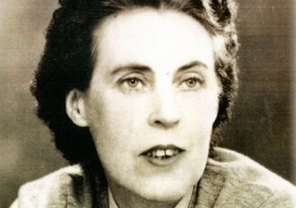 A black and white photograph of Una Troy, a woman with short, dark, wavy hair. She is wearing a light-colored top and looking slightly to the side with a neutral expression. The background is blurred, evoking the timeless essence of an author in contemplation.