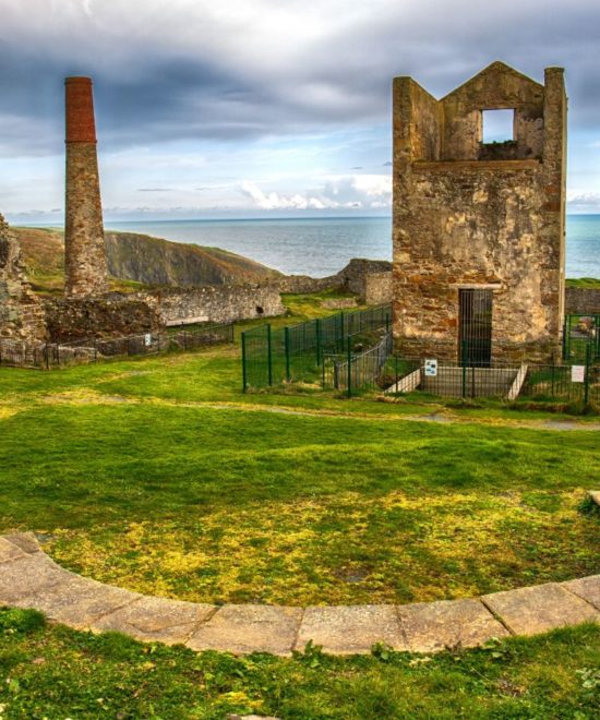 Ruins of an old industrial home with a brick chimney, overlooking a rugged coastline under a dramatic sky.