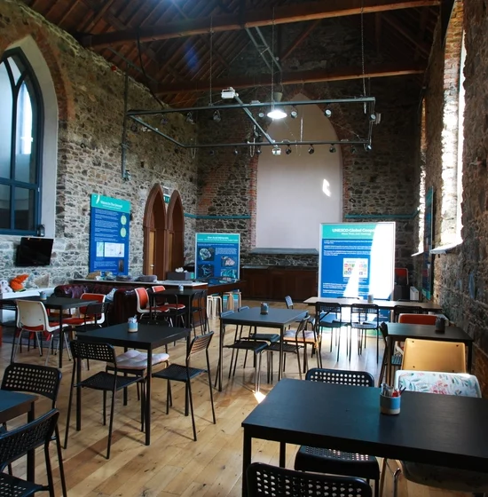 A spacious, well-lit room with rustic stone walls and a high wooden ceiling. The room, part of the Visitor Centre, is furnished with black tables and chairs positioned neatly. Large windows allow natural light in. Poster boards and information displays are set against the walls and near the front.