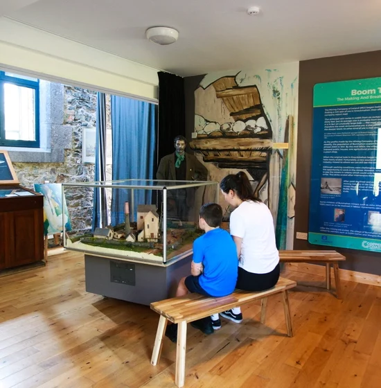 A woman and a boy sit on a wooden bench at the Visitor Centre, admiring a model display in the museum exhibit. The room features informational plaques on the walls, wooden display cases, and historical artifacts. A life-size mannequin stands in the background.