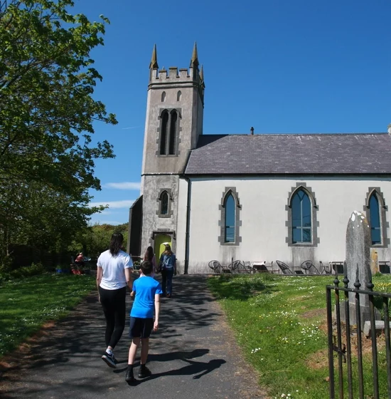 A group of visitors, including a man and a child, walk towards a small stone church with a prominent tower and arched windows. The church is surrounded by a grassy area with trees and other buildings visible in the background under the blue sky, creating an inviting centre for community gatherings.