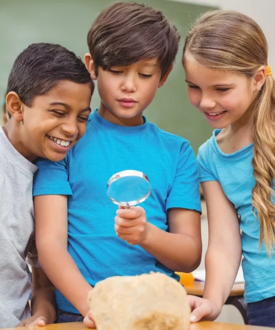 Three children are closely examining a rock with a magnifying glass. They are all smiling and seem engaged, participating in what appears to be a school programme. The children are wearing blue shirts and are in an indoor setting, likely a classroom. The background features a chalkboard and a desk.