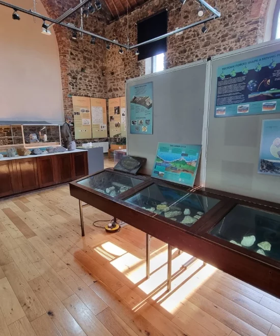 A museum exhibit room with stone walls and a wooden floor. Glass display cases with various artifacts are in the foreground, and information boards with text and images are mounted on partitions. Additional artifacts, curated to reflect the rich history of this UNESCO Global Geopark, are displayed in wooden cabinets along the wall.