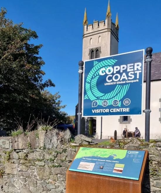 A stone building with a tall, pointed clock tower stands behind a large sign that reads "Copper Coast UNESCO Global Geopark Visitor Centre." In the foreground, there is an informational display panel on park management. Trees and a stone wall are visible on a bright sunny day.