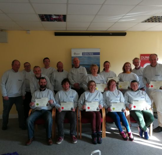 A group of people is posing indoors, all wearing grey sweatshirts. Some are standing and some are seated, holding certificates. Everyone is smiling at the camera. The room has beige walls, a poster, and a "DUN" visible in the background—celebrating with the Copper Coast Network.