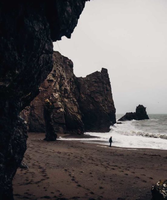 A person stands alone on a rocky beach with tall, rugged cliffs and crashing waves under a cloudy, overcast sky. The scene is viewed from within a dark cave, creating a frame around the coastal landscape. This dramatic setting offers an awe-inspiring glimpse into the fascinating world of geology.