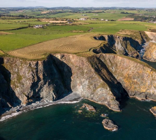 Aerial view of a rugged coastline with steep cliffs plunging into the sea. The top of the cliffs features lush green fields and farmland, revealing traces of mining heritage. The ocean below is a deep blue, with scattered rocks near the shore. Rolling hills extend into the horizon under a clear sky.